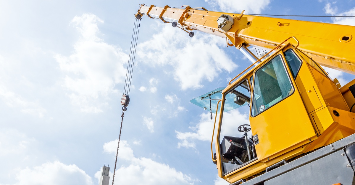 A yellow crane with an extended hook and chain against a bright sky, near concrete pillars at a construction site.