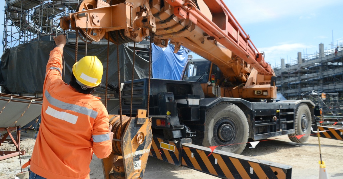 A worker wearing a safety jacket and a hard hat adjusts the hook block of a large crane at a construction site.